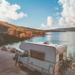person leaning against white RV and looking at a lake